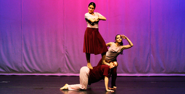 Três bailarinos no centro do palco, a figura masculina em posição de engatinho, onde uma das bailarinas está em pé nas costas dele e a terceira bailarina em posição de dança na parte de três