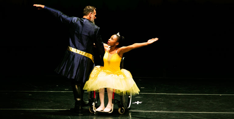 Foto de casal de bailarinos, onde a bailarina está de cadeira de rodas e estão performando uma dança.