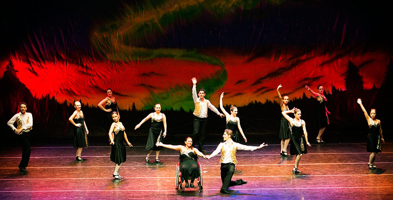 Grupo de bailarinos e bailarinas posicionados e distribuidos uniformemente no palco, com a apresentação em destaque de uma bailarina cadeirante
