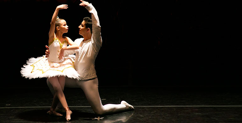 Imagem de um casal de bailarinos onde a parceira é uma menina sentada no bailarino que está em posição de cortejo