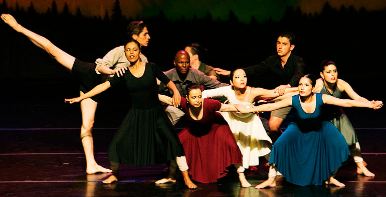 Nove bailarinos aglomerados no palco com braços abertos dramatizando uma situação ritmica.