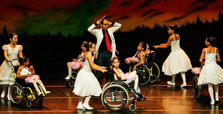 Cinco bailarinas cadeirante sendo conduzidas por outras cinco bailarinas, e no centro um homem em posição de aguardo.