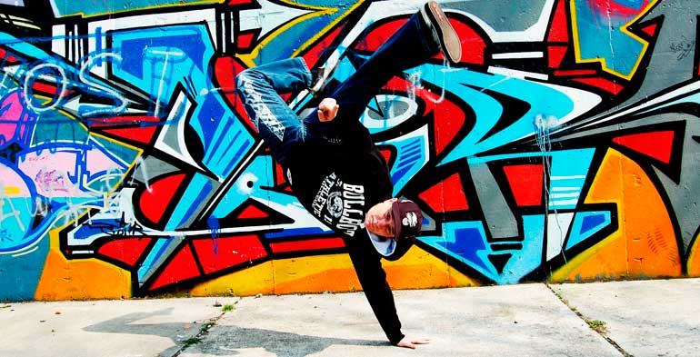 Um dançarino de hip hop em movimento radical, onde está apoiado apenas com uma mão de cabeça para baixo em um fundo todo grafitado e bem colorido.