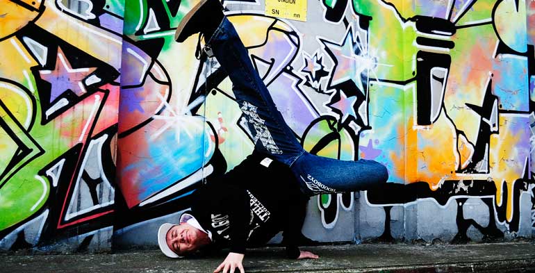 Dançarino de hip hop suspende seu corpo com as duas mãos em um fundo todo grafitado e bem colorido.