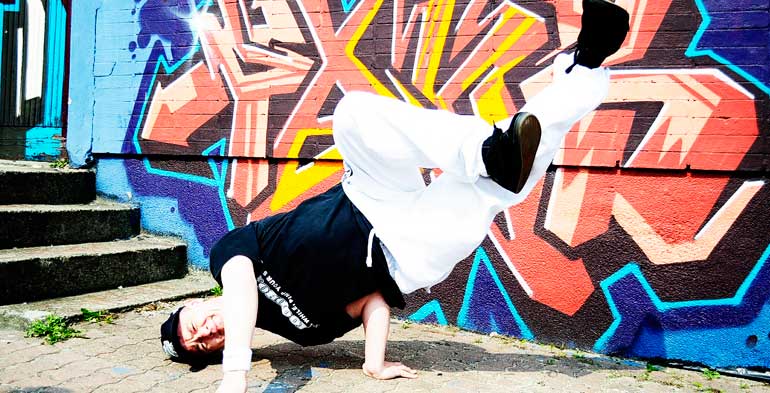 Dançarino de hip hop suspende seu corpo e cruz as pernas no ar com as duas mãos em um fundo todo grafitado e bem colorido
