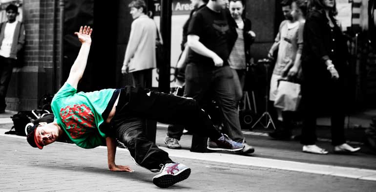 o dançarino de hip hop está girando o corpo com o apoio de apenas uma mão no chão, e na imagem total tem um tratamento gráfico onde tudo está em preto e branco, e só o dançarino está colorido.