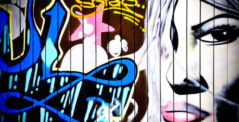 muro grafitado com a figura de uma mulher olhando fixamente e na frente dela a assinatura do artista desenhado com estilo e cores bem expressivos.